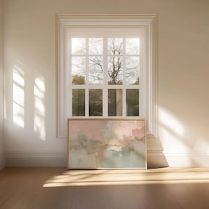 May include: An abstract painting in a wooden frame, featuring soft pastel colors of pink, beige, and blue. The artwork is displayed on a wooden floor, positioned beneath a white window with a view of a tree and greenery.