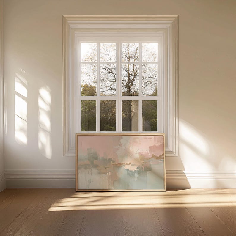 May include: An abstract painting in a wooden frame, featuring soft pastel colors of pink, beige, and blue. The artwork is displayed on a wooden floor, positioned beneath a white window with a view of a tree and greenery.