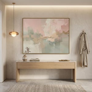 May include: An abstract painting in a gold frame hangs above a wooden console table. The painting features soft pink, green, and beige hues. A beige rug and a coat hanging on the wall complete the interior design.