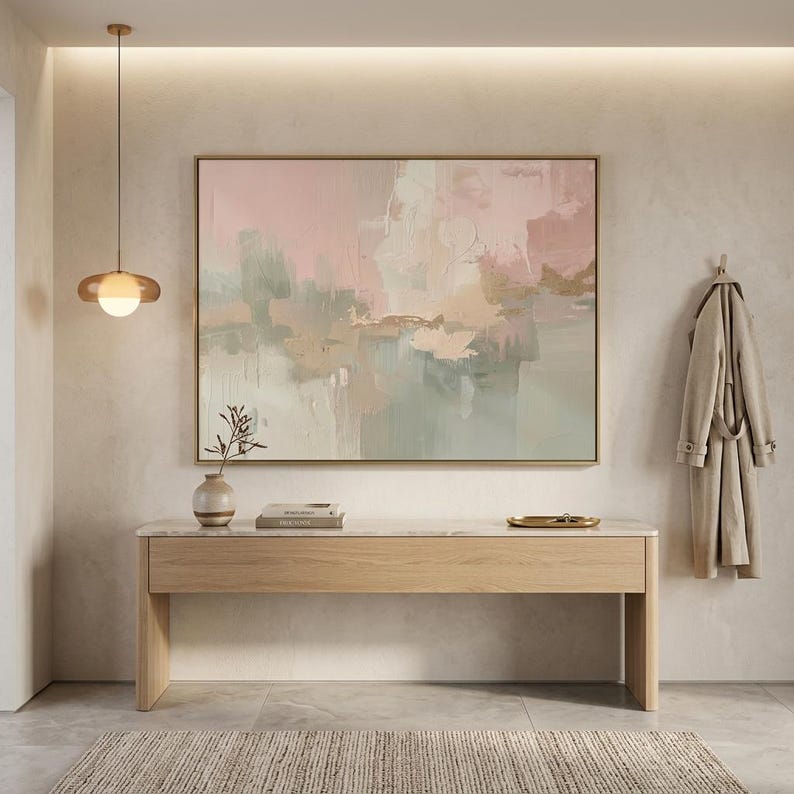 May include: An abstract painting in a gold frame hangs above a wooden console table. The painting features soft pink, green, and beige hues. A beige rug and a coat hanging on the wall complete the interior design.