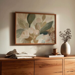 May include: A floral painting in a wooden frame hangs on a wall. The artwork features soft hues of green, pink, and beige. A wooden cabinet with drawers is below the painting. A vase with dried flowers and folded fabric are on the cabinet.