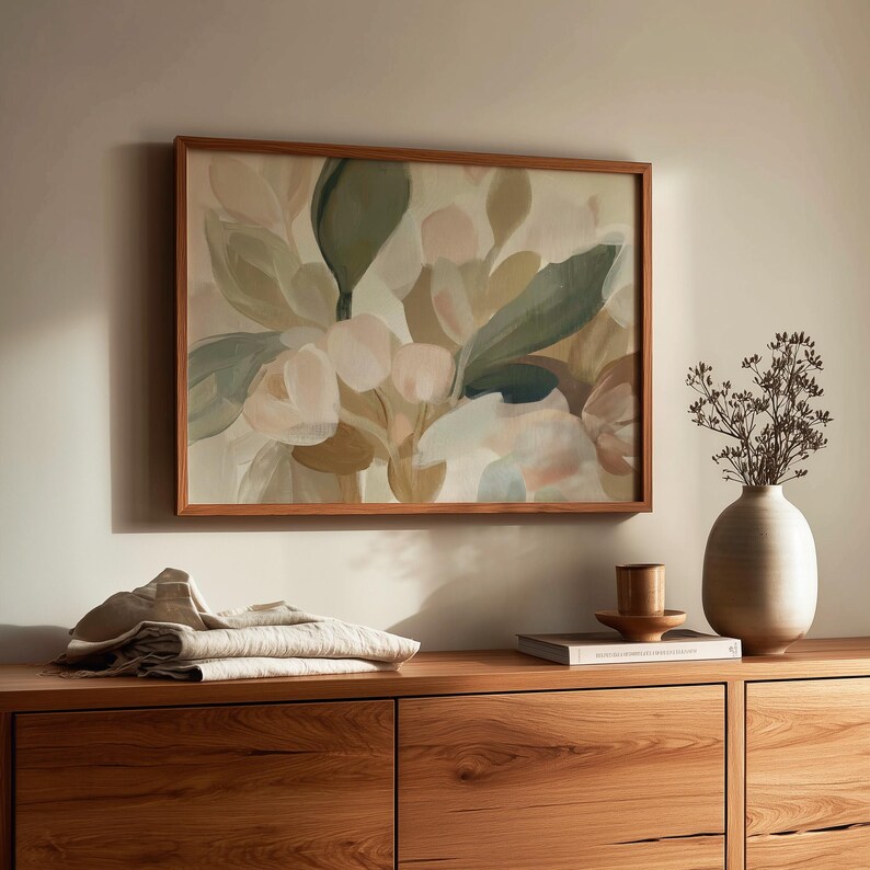 May include: A floral painting in a wooden frame hangs on a wall. The artwork features soft hues of green, pink, and beige. A wooden cabinet with drawers is below the painting. A vase with dried flowers and folded fabric are on the cabinet.
