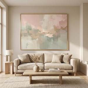 May include: Large abstract painting in a gold frame, featuring soft pink, beige, and muted green brushstrokes. The artwork hangs above a beige sofa with matching pillows, a wooden coffee table, and a textured rug. The room is bathed in natural light.