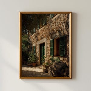 May include: A framed print of a stone building with green shutters and a green door. Potted plants and a stone path are in front of the building. The frame is gold.