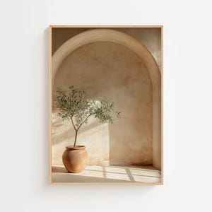 Op de afbeelding: Een ingelijste kunstdruk met een terracotta pot met een olijfboom in een gewelfde nis. De achtergrond is warm beige, met schaduwen door zonlicht. De lijst is licht van kleur.