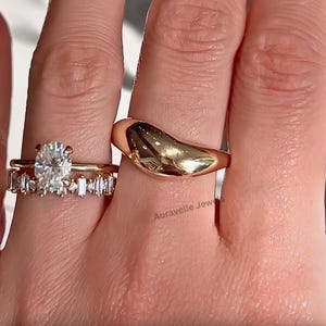 May include: A gold-toned ring with a smooth, domed surface and a stacked ring set featuring a central oval-cut stone flanked by baguette-cut stones. The text Auravelle Jewels is visible on the skin near the rings. The jewellery has a polished finish.