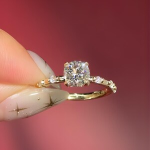 Puede incluir: Un anillo de oro con una gran piedra preciosa redonda y transparente como pieza central. La banda también es dorada y presenta piedras preciosas más pequeñas y transparentes. El anillo se sostiene sobre un fondo rojo.