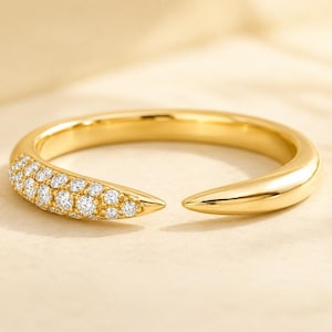 Könnte beinhalten: Ein offener Goldring mit spitz zulaufendem Design. Ein Ende des Rings ist mit mehreren kleinen, klaren, rund geschliffenen Steinen besetzt, während das andere Ende eine glatte, polierte Goldspitze bildet. Der Ring hat ein poliertes Finish.