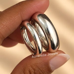 Puede incluir: Dos anillos plateados pulidos con un perfil liso, redondeado y abombado. Los anillos se sostienen juntos, mostrando una superficie metálica reflectante. El texto Auravelle Jewels está impreso en blanco en la parte inferior del anillo más grande.