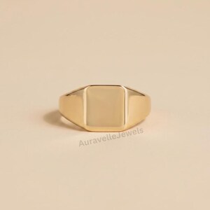 May include: A gold-toned signet ring featuring a smooth, flat, rectangular face and a polished band. The ring has a clean, solid construction with a reflective surface. The text AuravelleJewels is visible in small, dark letters below the ring.