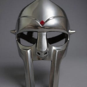 May include: A metallic silver-colored face mask featuring a smooth, polished finish with a pointed forehead section. A small, square-shaped red gemstone is embedded at the center of the brow. The mask includes wide eye openings and two vertical bars.