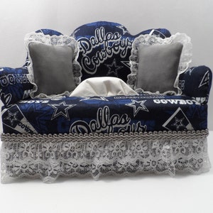 May include: A blue and white tissue box cover shaped like a couch with a Dallas Cowboys theme. The couch has two grey pillows and a white tissue box inside. The couch has a lace trim and a silver trim.