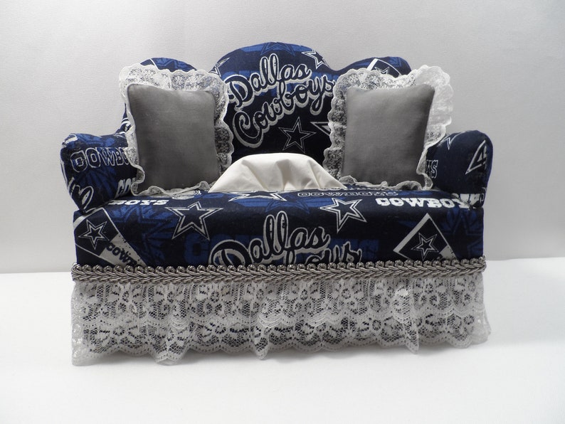 May include: A blue and white tissue box cover shaped like a couch with a Dallas Cowboys theme. The couch has two grey pillows and a white tissue box inside. The couch has a lace trim and a silver trim.