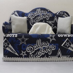 May include: A blue and white tissue box cover shaped like a couch with Dallas Cowboys logos and stars. The couch has two white cushions and a white tissue box.