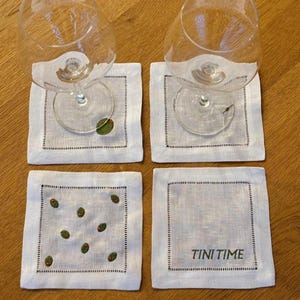 May include: Four white linen coasters with a black stitched border. Two coasters have a single embroidered green olive, one has multiple olives, and one reads "TINI TIME" in black text. Two wine glasses rest on the coasters.