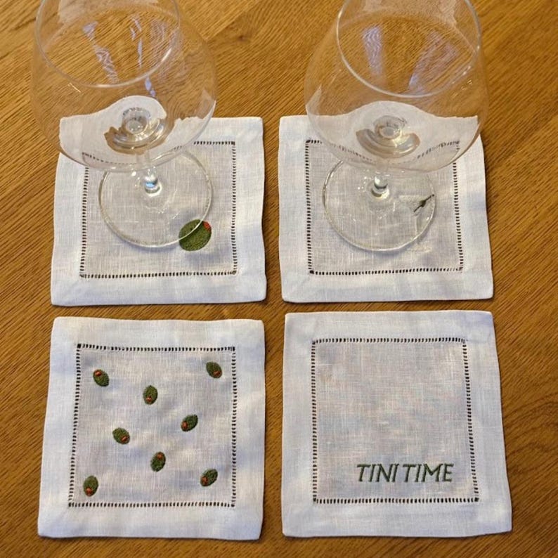 May include: Four white linen coasters with a black stitched border. Two coasters have a single embroidered green olive, one has multiple olives, and one reads "TINI TIME" in black text. Two wine glasses rest on the coasters.
