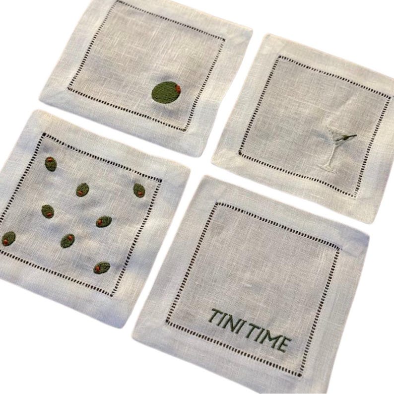 May include: Four square, light gray linen coasters with black stitched borders. Two coasters feature embroidered green olives, one has a martini glass, and another reads "TINI TIME" in green thread. A stylish set for entertaining.