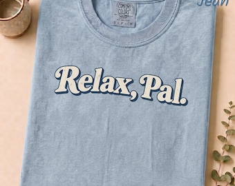 May include: A blue jean coloured short-sleeve t-shirt with a crew neckline. The shirt features the text Relax, Pal. printed in a cream colour with a dark blue outline across the chest. A Comfort Colors brand label is visible at the inner neck.