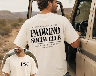 May include: An ivory short-sleeve t-shirt featuring black text. The back displays PROUD MEMBER OF THE PADRINO SOCIAL CLUB, PADRINO DE MATEO, and JUNE 14, 2026. The front chest area features the letters PSC and the text PADRINO SOCIAL CLUB.