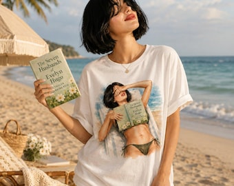 May include: A white short-sleeved t-shirt featuring a graphic print of a person in a bikini holding a book titled The Seven Husbands of Evelyn Hugo by Taylor Jenkins Reid. The graphic shows a beach scene with blue water and sand.