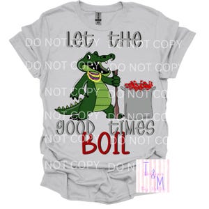 May include: A light grey t-shirt with a cartoon alligator wearing a purple and gold necklace, holding a wooden spoon, and standing next to a pot of red crawfish. The text reads "Let the good times BOIL."