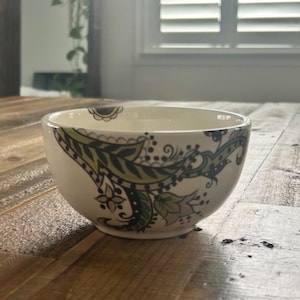 May include: A white ceramic bowl with a decorative paisley design in shades of green and gray. The bowl has a rounded shape and sits on a wooden surface. The design features floral and leaf motifs.