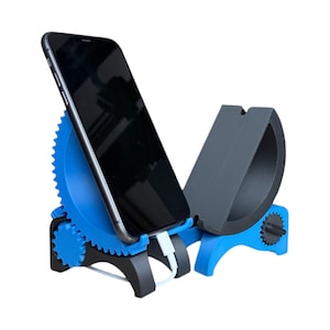 Può includere: Un supporto per telefono blu e nero con un design a forma di ingranaggio. Il supporto contiene uno smartphone nero, con un cavo di ricarica bianco collegato. Il supporto ha una seconda sezione nera per contenere un altro dispositivo.
