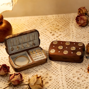 May include: A wooden jewelry box with an open compartment displaying gold necklaces, rings, and a bracelet. The closed box features gold rose designs and small white pearls. Dried roses are scattered around.