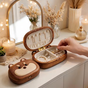 May include: Two oval wooden jewellery boxes with a dark brown finish and a light-coloured bear face design on the lids. One box is open, revealing a beige fabric interior with compartments for rings, necklaces, and bracelets, while the other is closed.