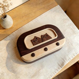 Puede incluir: Una caja de madera con un diseño de radio retro. La caja presenta un marco marrón oscuro y un centro de madera clara. Dos siluetas de gatos están talladas en la parte superior. Tres pequeños botones redondos de color marrón oscuro están en la parte delantera.