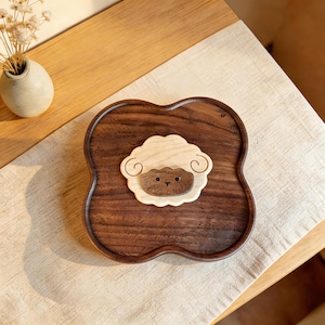 Pode incluir: Uma bandeja de madeira castanha escura em forma de trevo de quatro folhas. Apresenta um desenho de ovelha castanha clara no centro. A ovelha tem um corpo lanoso castanho claro e uma cara castanha escura. A bandeja mede cerca de 20 cm de diâmetro.