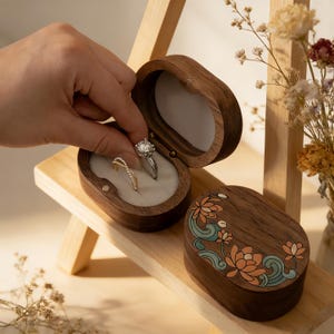 以下が含まれることがあります： 蓋に花の模様が入った楕円形の木製リングボックス。開くと2つの指輪が見える。ボックスの内側は明るい色。指輪はダイヤモンドの婚約指輪とゴールドバンド。