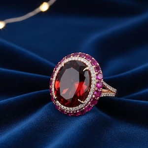 May include: An oval-shaped ring with a large, faceted, deep red gemstone as the centerpiece. The gemstone is surrounded by a halo of smaller, pink gemstones and a row of clear, sparkling stones. The ring has a gold band and is set against a dark blue background.