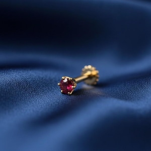 Könnte beinhalten: Ein einzelner goldener Ohrring mit einem kleinen, runden, tiefroten Edelstein. Der Ohrring ist vor einem Hintergrund aus tiefblauem Stoff platziert, der die Farbe und das Design des Schmucks hervorhebt. Der Ohrringstift hat eine strukturierte, dekorative Rückseite.