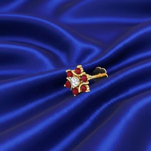 Może przedstawiać: Złoty kolczyk do nosa z kwiatowym wzorem z czerwonymi kamieniami szlachetnymi i centralnym diamentem. Biżuteria jest prezentowana na niebieskim satynowym tle.
