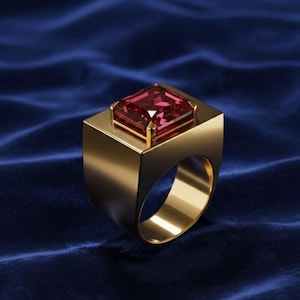 May include: A gold-colored ring with a large, square, red gemstone. The ring has a thick band and a square setting that holds the gemstone. The background is a dark blue fabric.