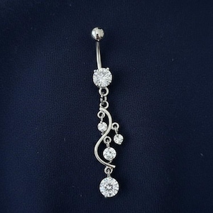 May include: A silver belly button ring with a curved design. The ring features a large, round, clear gemstone at the bottom and top, with smaller gemstones dangling from a curved silver bar. The background is a dark blue.