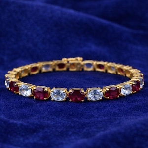 Peut inclure: Un bracelet en or orné de pierres précieuses ovales rouges et bleu clair alternées. Le bracelet est présenté sur un fond bleu foncé, mettant en valeur le design du bijou. Le fermoir est visible.