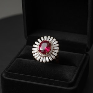 May include: A ruby and diamond ring set in a black velvet jewelry box. The ring features a large, oval-cut ruby surrounded by a halo of baguette-cut diamonds. The ring's band is gold-colored.