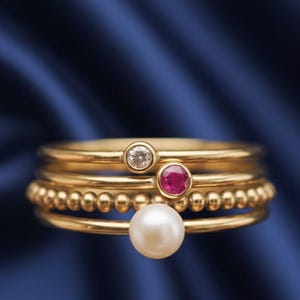 Peut inclure: Plusieurs bagues dorées superposées. L'une est ornée d'une pierre précieuse transparente, une autre d'une pierre rouge, une troisième présente une texture perlée, et la dernière une grosse perle blanche. Fond bleu foncé.