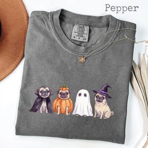 May include: A charcoal grey crew neck t-shirt featuring a graphic of four pugs in Halloween costumes: a vampire, a pumpkin, a ghost, and a witch. The shirt has a Comfort Colors label at the neck and the word Pepper is printed in the top right corner.