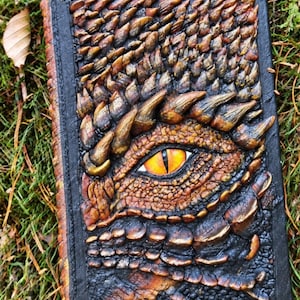Peut inclure: Un carnet rectangulaire avec une couverture texturée représentant un œil de dragon en relief dans des tons orange et jaune. La surface est sculptée d'écailles brunes et noires, avec quatre rivets métalliques aux coins sur un bord noir.