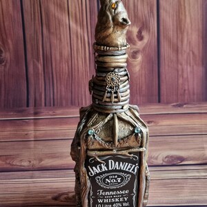 Peut inclure: Une bouteille de whisky Jack Daniel's décorative avec un dessus en forme de tête de loup. La bouteille est enfermée dans une sculpture détaillée aux tons métalliques avec des ornements. L'étiquette indique "Jack Daniel's Old No. 7 Tennessee Whiskey".