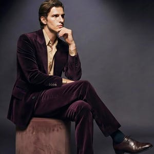 May include: A man in a plum velvet suit and a gold-colored shirt sits on a pink ottoman with a gold base. The word "VEMIRA" is displayed above the man. He wears brown leather dress shoes.