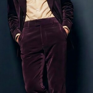 May include: A burgundy velvet suit, consisting of a blazer and trousers, paired with a light tan button-down shirt. The suit is a deep, rich color, and the outfit is completed with brown leather dress shoes.