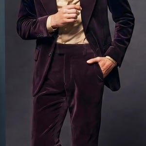 May include: A dark burgundy velvet suit with a matching blazer and trousers. The suit is paired with a light tan button-down shirt. The model is wearing brown leather dress shoes.