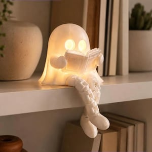 Könnte beinhalten: Eine weiße, 3D-gedruckte Geisterfigur mit leuchtenden, kreisförmigen Augen, die auf einem Regal sitzt und ein offenes Buch hält. Der Geist hat bewegliche, baumelnde Beine und wird von innen beleuchtet. Der Text BUNDLE STL + 3MF ist sichtbar.