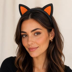 May include: A black headband featuring two upright cat ears. Each ear has a solid black outer border and a vibrant orange triangular center. The headband is constructed with a smooth, matte finish and is designed to sit securely on the head.