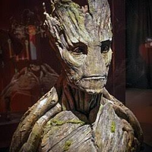 May include: A detailed bust of a humanoid figure with a bark-like texture, featuring deep brown and grey wood grain patterns, patches of green moss, and distinct facial features including eyes, a nose, and a mouth, resembling a tree-like creature.