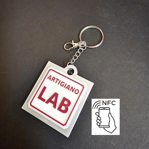 Könnte beinhalten: Ein quadratischer Schlüsselanhänger mit weißer Basis, rotem Rand und der Aufschrift ARTIGIANO LAB in roten Buchstaben. Der Anhänger verfügt über einen silbernen Metallring und einen Karabinerverschluss. Ein NFC-Symbol mit einer Hand am Telefon ist zu sehen.